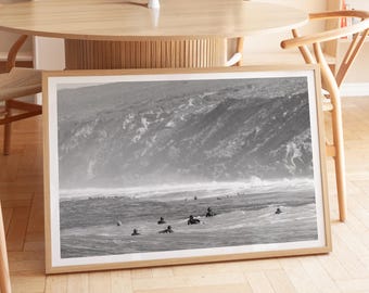 May include: A framed black and white photograph of surfers in the ocean. The image captures the surfers riding waves near a rocky coastline. The photograph is displayed in a light wood frame, leaning against a wooden chair and table.