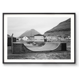 May include: A black and white photograph of a concrete half-pipe skate ramp in a grassy area. The ramp is in front of a building with a mountain in the background.
