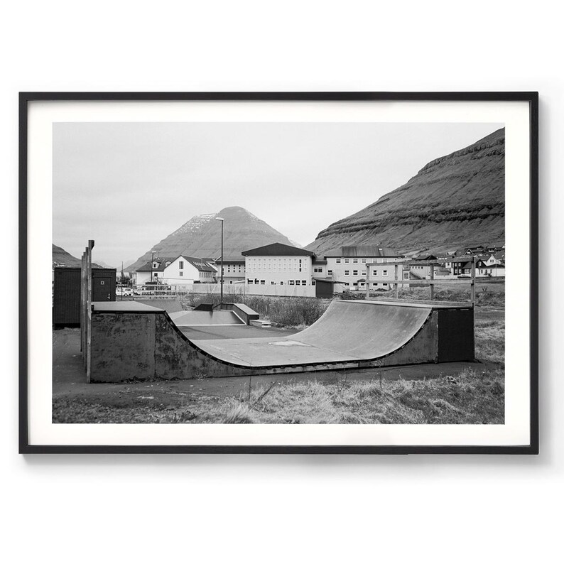 May include: A black and white photograph of a concrete half-pipe skate ramp in a grassy area. The ramp is in front of a building with a mountain in the background.