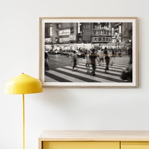 May include: Black and white framed photograph of a busy city street scene. People are crossing a crosswalk, and buildings and signs are visible in the background. The image has a blurred effect.