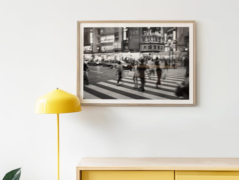 May include: Black and white framed photograph of a busy city street scene. People are crossing a crosswalk, and buildings and signs are visible in the background. The image has a blurred effect.