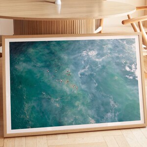 May include: A framed art print featuring an aerial view of the ocean. The artwork showcases turquoise and blue water with white foamy waves. Several dark figures are scattered across the water's surface, possibly surfers or swimmers.