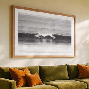May include: A large framed black and white photograph of surfers riding waves. The photograph is displayed above a green velvet sofa with orange throw pillows. The frame is a light brown color.