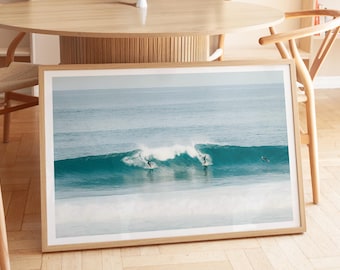 May include: A rectangular framed wall art print featuring a horizontal ocean scene with three surfers riding turquoise waves. The photograph displays a breaking wave with white foam, deep blue water, and a light sky, enclosed in a light wood frame.