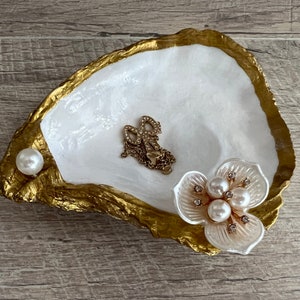 May include: A white and gold oyster shell dish with a pearl and a white flower with pearl accents. The dish is perfect for holding jewelry or other small items.