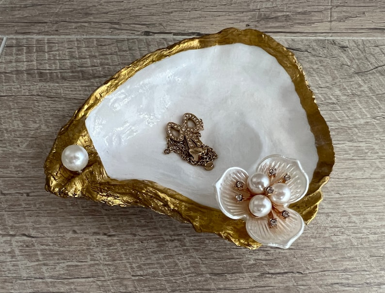 May include: A white and gold oyster shell dish with a pearl and a white flower with pearl accents. The dish is perfect for holding jewelry or other small items.