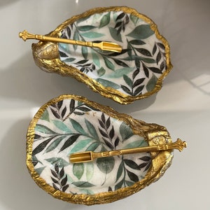 Könnte beinhalten: Zwei kleine, weiße Keramik-Austernmuscheln mit goldenen Verzierungen. Jede Muschel hat ein grünes und schwarzes Blattmotiv und enthält einen goldfarbenen Löffel.
