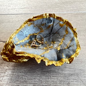 May include: A grey concrete ring dish with a gold leaf design. The dish is shaped like a seashell and has a small indentation in the centre.