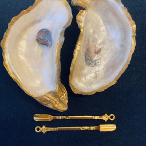 May include: Two oyster shells painted gold, with a pair of gold-toned metal spatulas. The spatulas have a decorative design on the handles.
