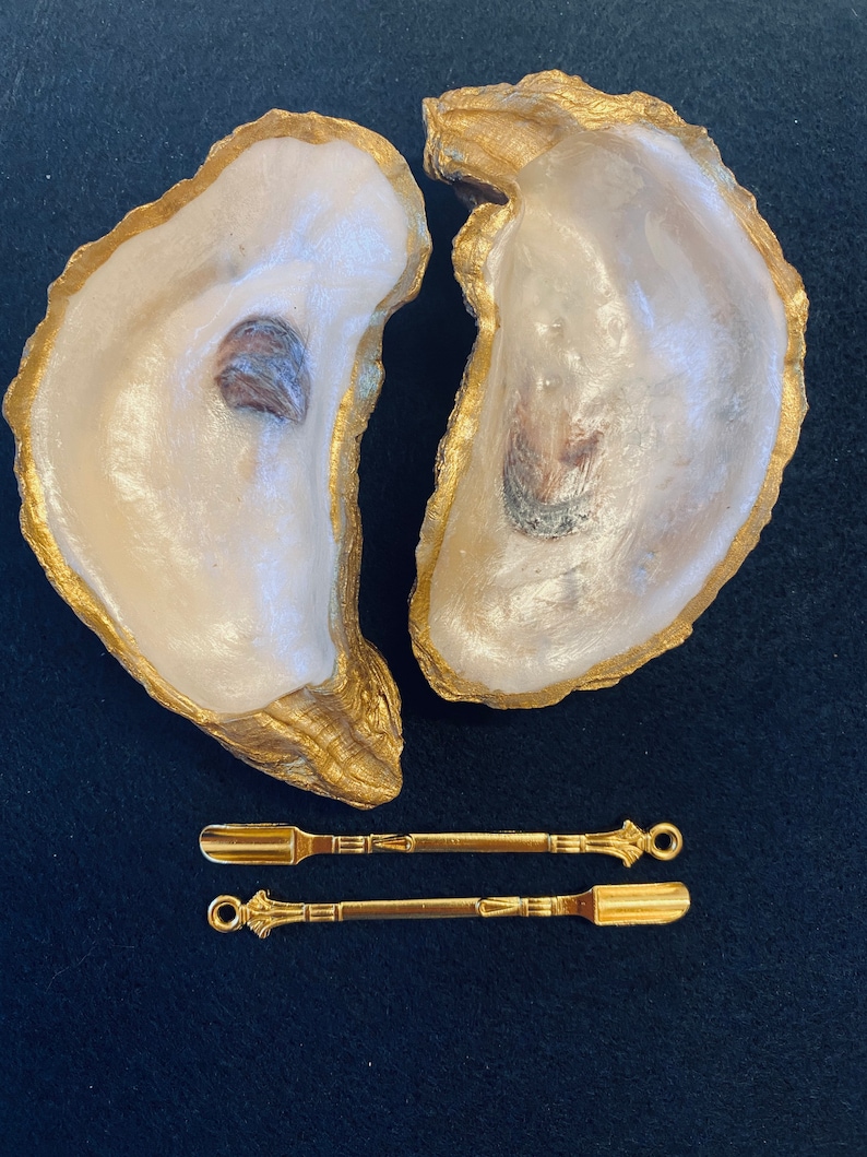 May include: Two oyster shells painted gold, with a pair of gold-toned metal spatulas. The spatulas have a decorative design on the handles.