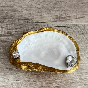 Pode incluir: Um prato de concha de ostra branca com borda dourada e dois acentos de strass transparentes.