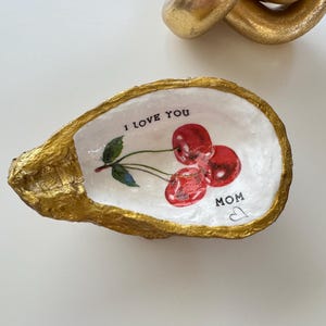 Könnte beinhalten: Eine weiße Austernmuschel-Schale mit goldener Verzierung, die mit einem gemalten Design aus roten Kirschen und dem Text "I love you Mom" mit einem Herzen verziert ist.