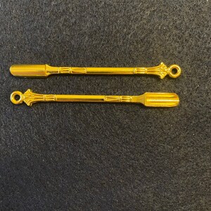 May include: Two gold metal scoops with ornate handles. The scoops are approximately 10 cm long and have a flat, rectangular shape.