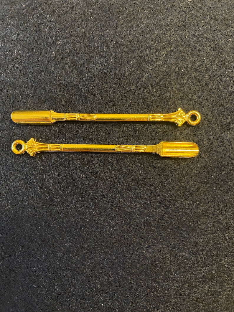 May include: Two gold metal scoops with ornate handles. The scoops are approximately 10 cm long and have a flat, rectangular shape.