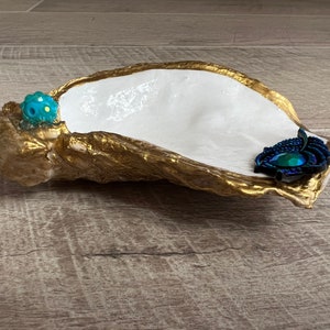 May include: A white oyster shell dish with gold accents. The dish has a blue and green jewel and a blue and black feather accent.