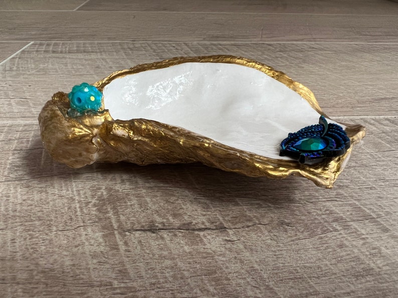 May include: A white oyster shell dish with gold accents. The dish has a blue and green jewel and a blue and black feather accent.