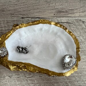 Pode incluir: Um prato de concha de ostra branca com borda dourada, usado como bandeja para joias. O prato est&aacute; cheio de tr&ecirc;s pe&ccedil;as de joias de strass transparentes.