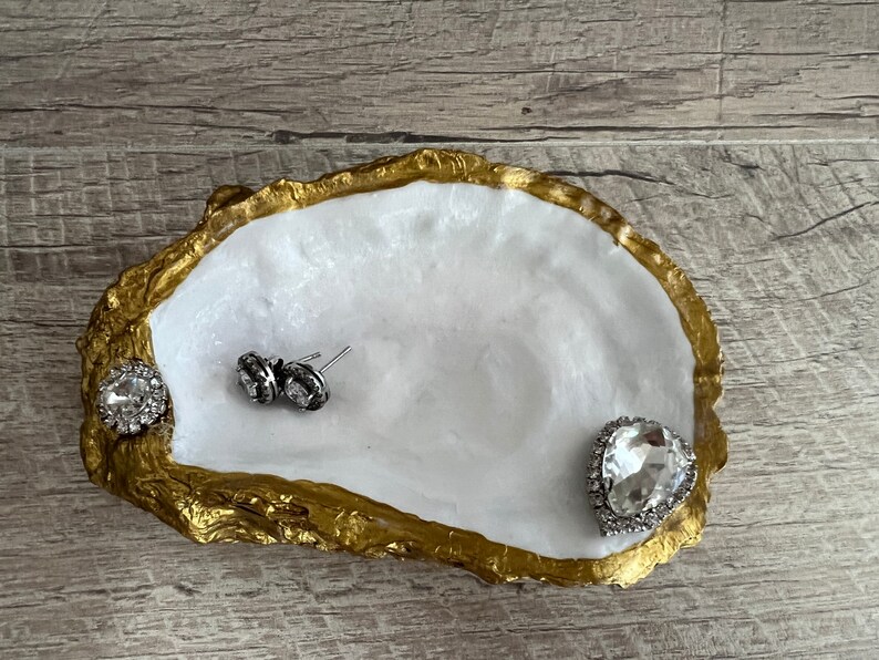 Pode incluir: Um prato de concha de ostra branca com borda dourada, usado como bandeja para joias. O prato est&aacute; cheio de tr&ecirc;s pe&ccedil;as de joias de strass transparentes.