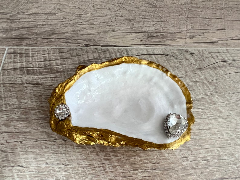 Pode incluir: Um prato de concha de ostra branca com borda dourada. O prato tem dois enfeites de strass transparentes.