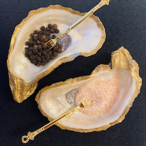 May include: Two gold oyster shell salt and pepper shakers with matching gold spoons. The oyster shells are white with a pearlescent finish. One shell contains black peppercorns and the other contains pink Himalayan salt.