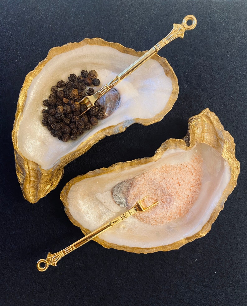 May include: Two gold oyster shell salt and pepper shakers with matching gold spoons. The oyster shells are white with a pearlescent finish. One shell contains black peppercorns and the other contains pink Himalayan salt.