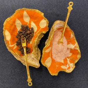 Puede incluir: Dos saleros y pimenteros de cerámica naranja con detalles dorados. Los saleros tienen forma de concha y presentan un diseño floral blanco. Un salero está lleno de granos de pimienta negra y el otro de sal rosa. Se incluyen dos cucharas doradas.