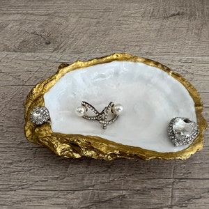 Pode incluir: Um prato de concha de ostra branca com detalhes dourados, usado como porta-joias. Apresenta um pequeno anel de cora&ccedil;&atilde;o prateado, um par de brincos de p&eacute;rola e um anel de prata com um design de estrela.