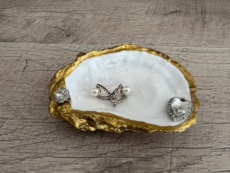 Pode incluir: Um prato de concha de ostra branca com detalhes dourados, usado como porta-joias. Apresenta um pequeno anel de cora&ccedil;&atilde;o prateado, um par de brincos de p&eacute;rola e um anel de prata com um design de estrela.