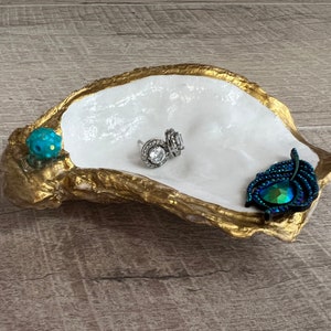 May include: A white oyster shell dish with gold accents, decorated with a blue and green jewel and a peacock feather. The dish is perfect for holding jewelry or other small items.