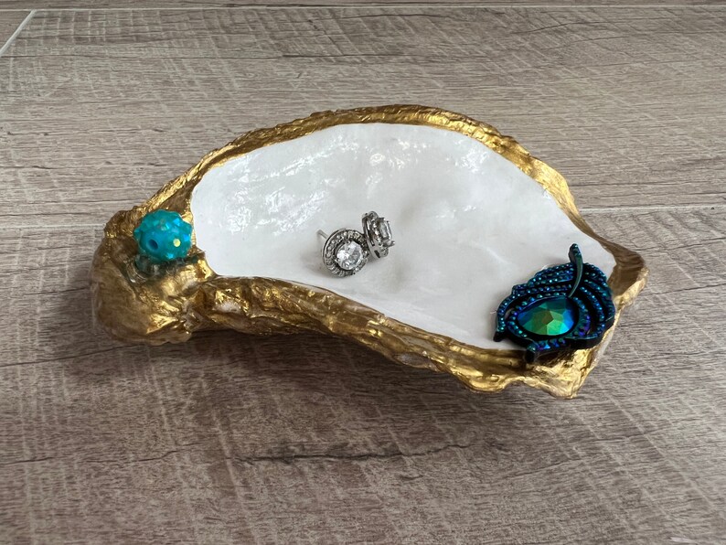 May include: A white oyster shell dish with gold accents, decorated with a blue and green jewel and a peacock feather. The dish is perfect for holding jewelry or other small items.