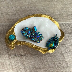 May include: A white oyster shell dish with gold trim, used as a jewelry holder. The dish contains three pieces of jewelry: a pair of blue and green iridescent earrings and a blue and green iridescent feather-shaped brooch.