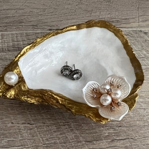 May include: A white oyster shell dish with gold accents, featuring a pearl and a flower with pearls and rhinestones. The dish is perfect for holding jewelry or other small items.