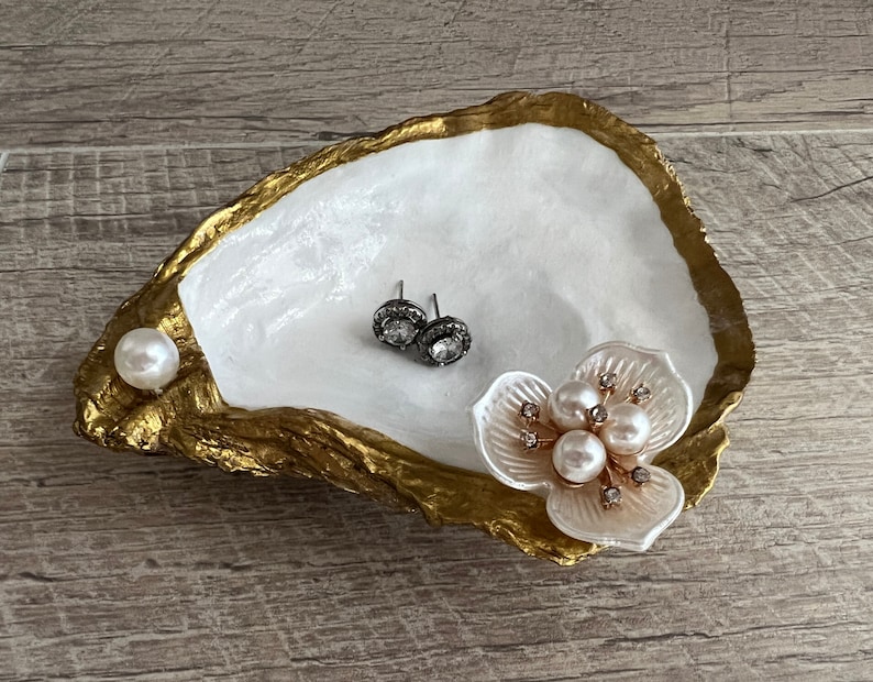 May include: A white oyster shell dish with gold accents, featuring a pearl and a flower with pearls and rhinestones. The dish is perfect for holding jewelry or other small items.