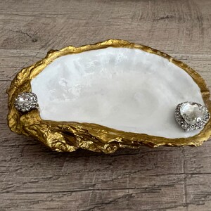 Pode incluir: Um prato de concha de ostra branca com detalhes dourados, adornado com dois strass transparentes. O prato &eacute; perfeito para guardar joias ou outros objetos pequenos.