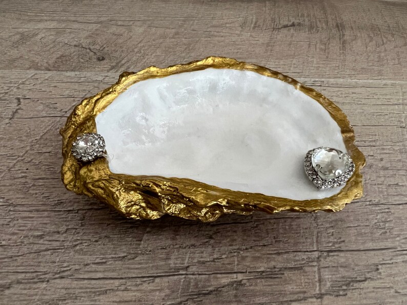 Pode incluir: Um prato de concha de ostra branca com detalhes dourados, adornado com dois strass transparentes. O prato &eacute; perfeito para guardar joias ou outros objetos pequenos.