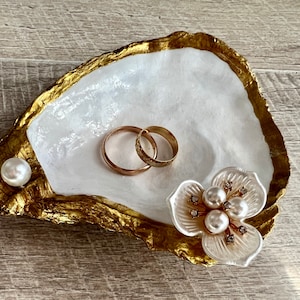 May include: A white oyster shell ring dish with gold accents. The dish features a pearl and a decorative flower with pearls and rhinestones.