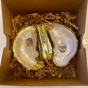 May include: Two white oyster shells with gold accents, used as dishes, are filled with brown paper packing material. A gold metal tool with a clear plastic handle is in the centre of the shells.