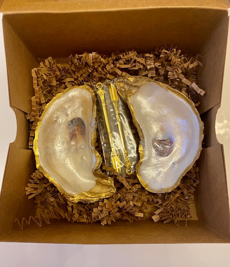 May include: Two white oyster shells with gold accents, used as dishes, are filled with brown paper packing material. A gold metal tool with a clear plastic handle is in the centre of the shells.