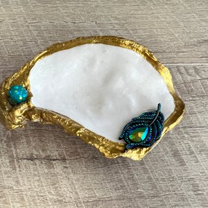 May include: A white oyster shell dish with a gold rim, decorated with a teal blue iridescent feather and a small teal blue gem.