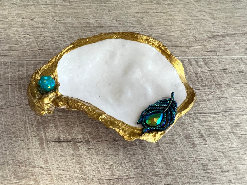 May include: A white oyster shell dish with a gold rim, decorated with a teal blue iridescent feather and a small teal blue gem.
