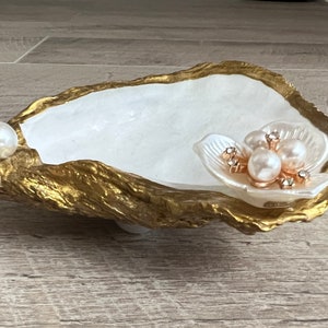 May include: A white oyster shell dish with a gold rim, decorated with a pearl and a flower with pearl accents.