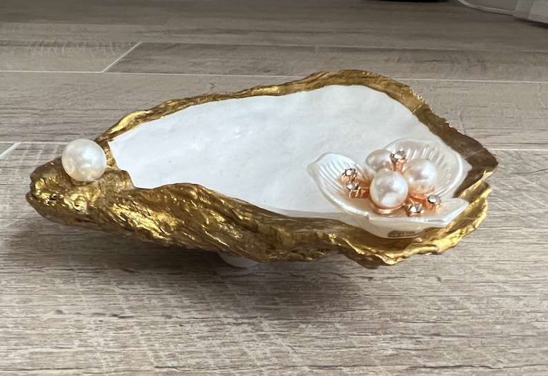 May include: A white oyster shell dish with a gold rim, decorated with a pearl and a flower with pearl accents.
