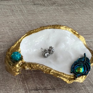 May include: A white oyster shell dish with gold accents, featuring a teal blue rhinestone, a pair of silver earrings with clear rhinestones, and a teal blue feather-shaped brooch with iridescent rhinestones.