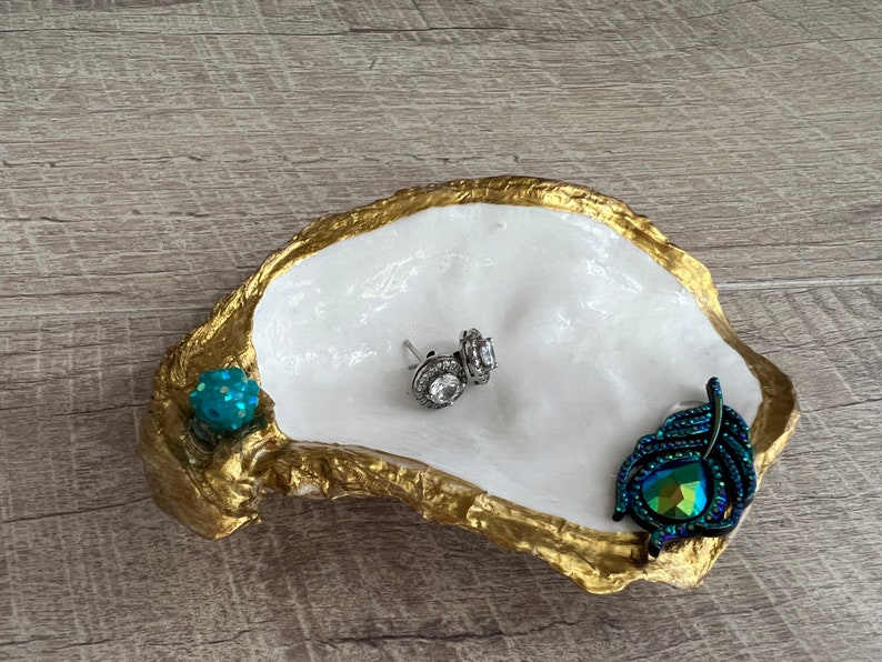 May include: A white oyster shell dish with gold accents, featuring a teal blue rhinestone, a pair of silver earrings with clear rhinestones, and a teal blue feather-shaped brooch with iridescent rhinestones.