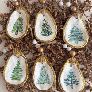 Puede incluir: Seis adornos navideños hechos a mano. Cada adorno es una concha de ostra con un borde dorado y un diseño de árbol de Navidad pintado. Los adornos son blancos con árboles verdes y azules. Cada adorno tiene una cuerda de yute para colgar.