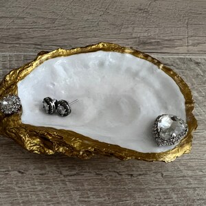 Pode incluir: Um prato de concha de ostra branca com borda dourada, usado como bandeja para joias. O prato tem tr&ecirc;s strass, dois pequenos redondos e um grande em forma de cora&ccedil;&atilde;o.