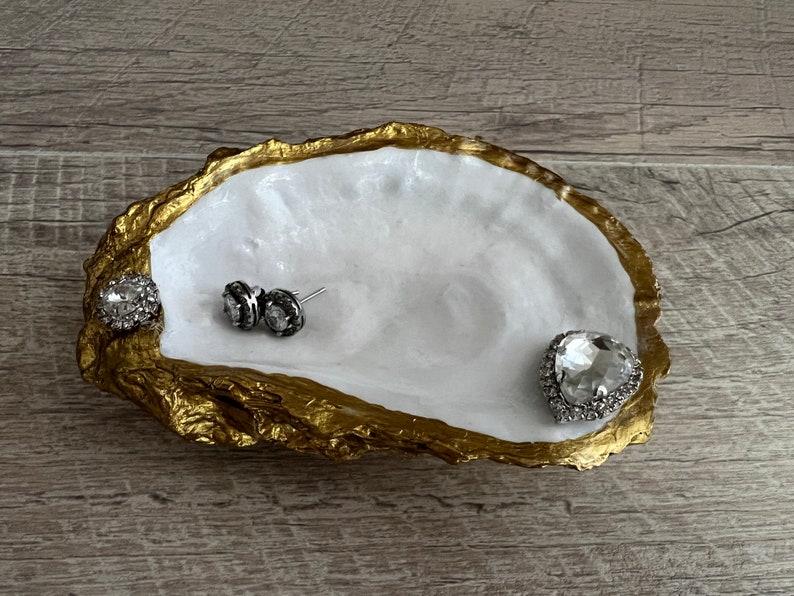 Pode incluir: Um prato de concha de ostra branca com borda dourada, usado como bandeja para joias. O prato tem tr&ecirc;s strass, dois pequenos redondos e um grande em forma de cora&ccedil;&atilde;o.
