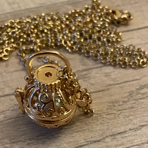 Puede incluir: Un collar de tono dorado con una cadena delicada y un colgante redondo en forma de jaula adornada. La jaula está abierta y tiene una pequeña abertura redonda en la parte superior.