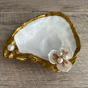 May include: A white oyster shell dish with gold accents, featuring a pearl and a white flower with pearl and rhinestone embellishments.
