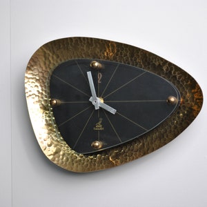 Può includere: Un orologio da parete in metallo martellato color oro con un quadrante nero e lancette argentate. Il quadrante dell'orologio ha un logo di uccello nero con il testo "JAY  HAMMOND" sotto.
